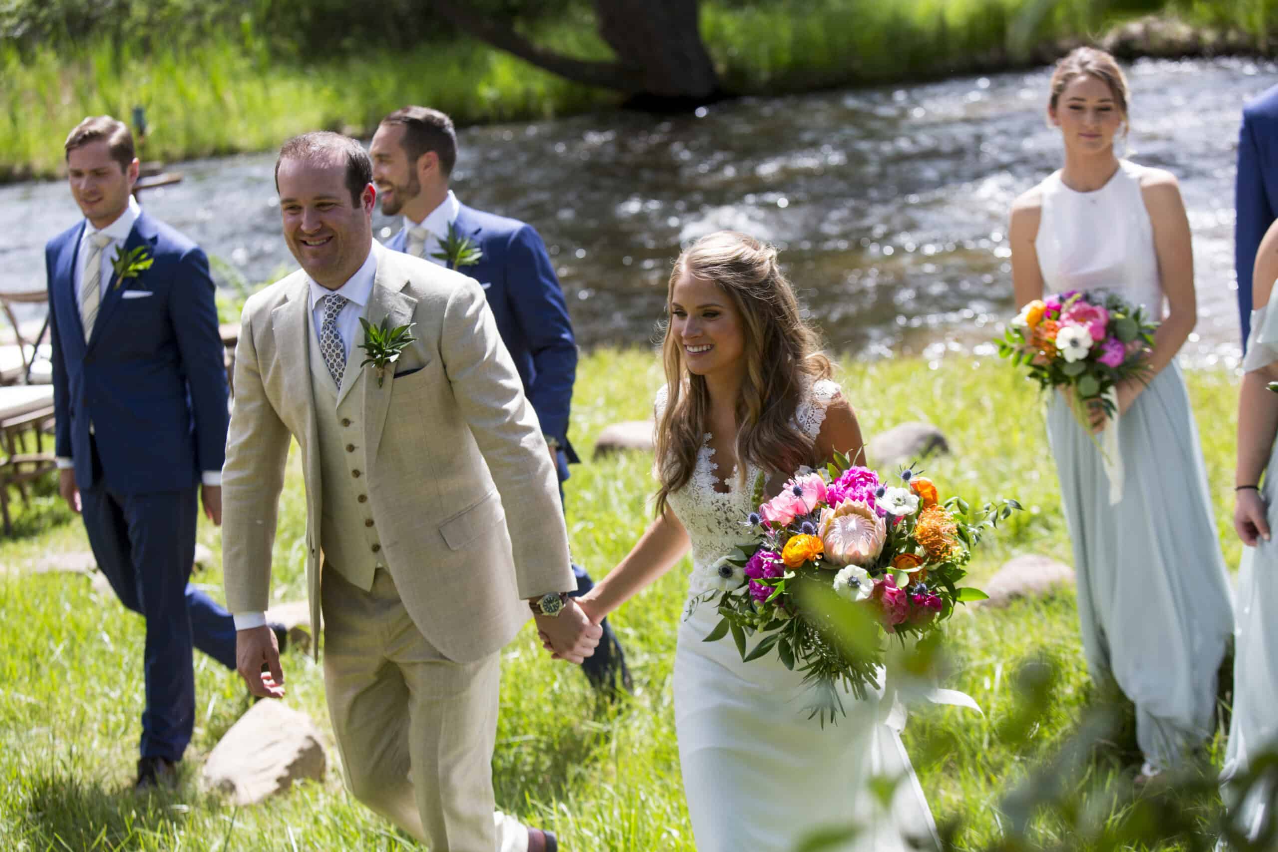 Bride and groom walking hand in hand with wedding party outdoors by a river.