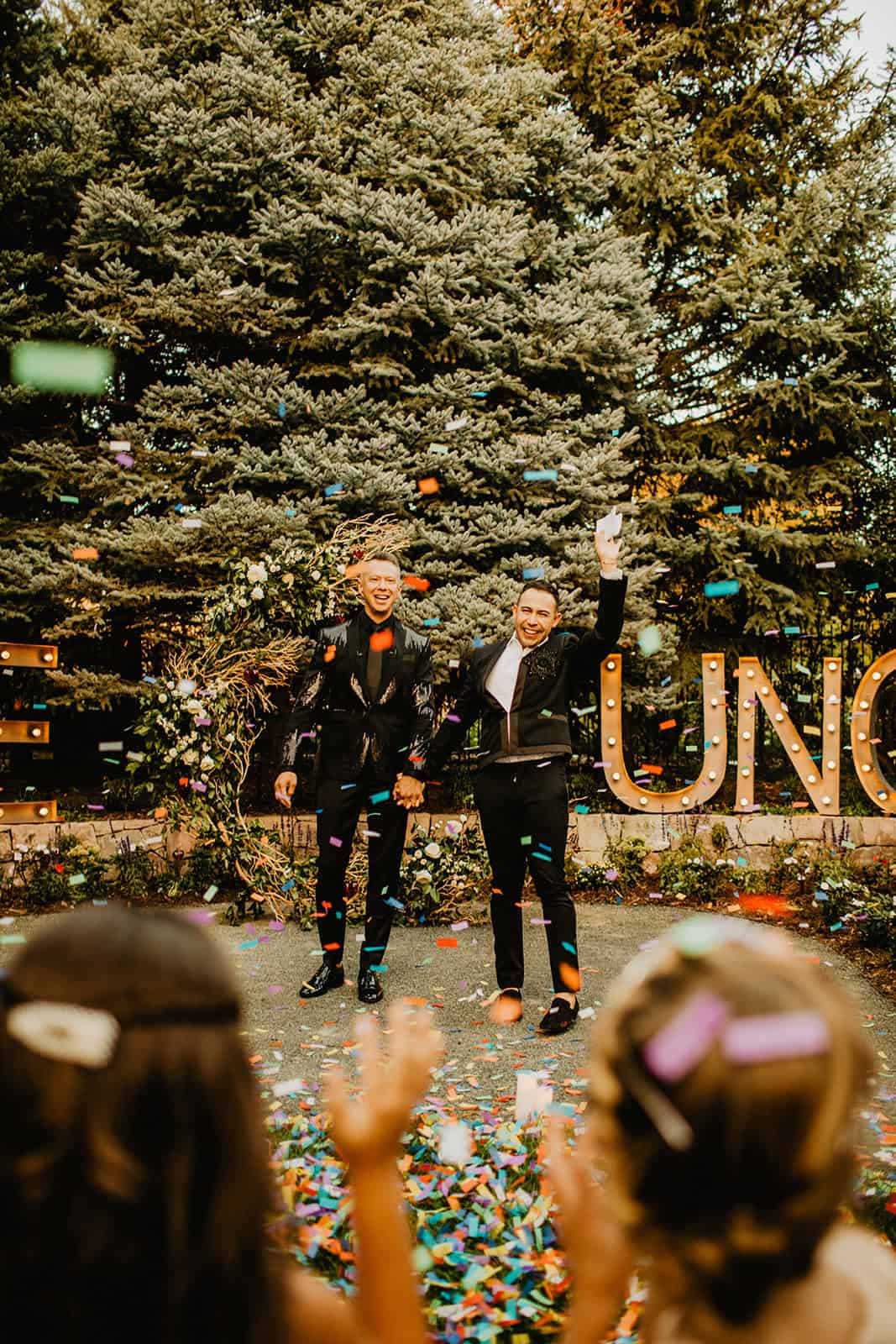 Two men celebrating at a wedding reception with confetti and a decorated outdoor setting.