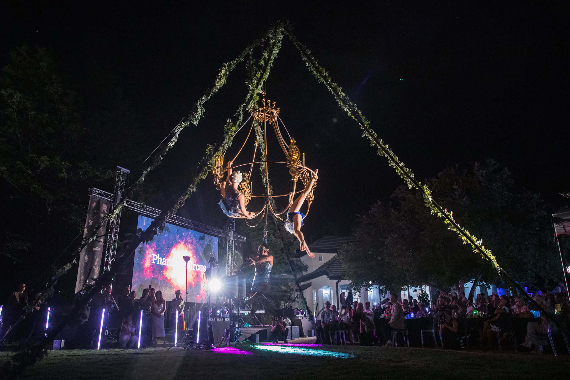 Aerial acrobats perform on a large hanging apparatus at night during a charity event.