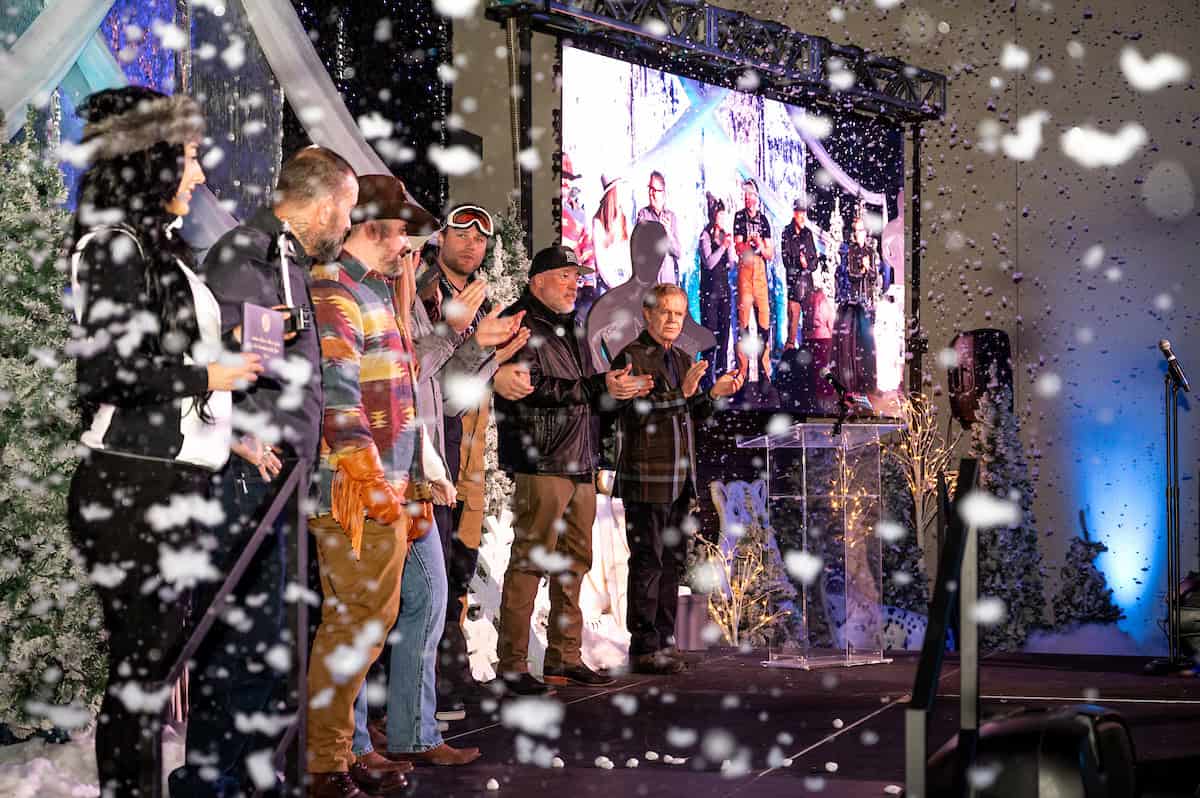 Group of people on stage at Winter Wonderland Gala with large screen and snow effects.