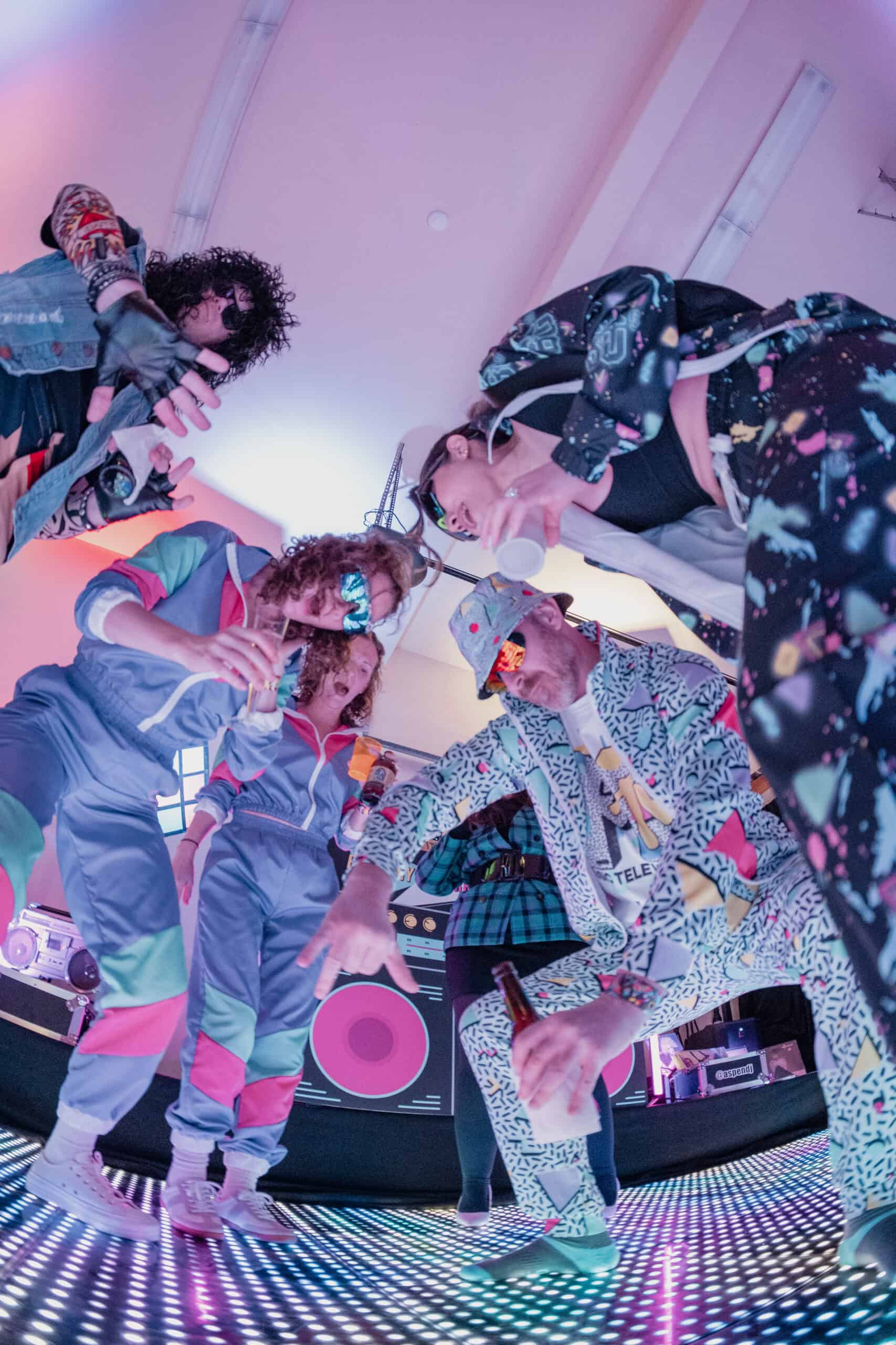 People dancing at a colorful party with vibrant lighting and DJ equipment.