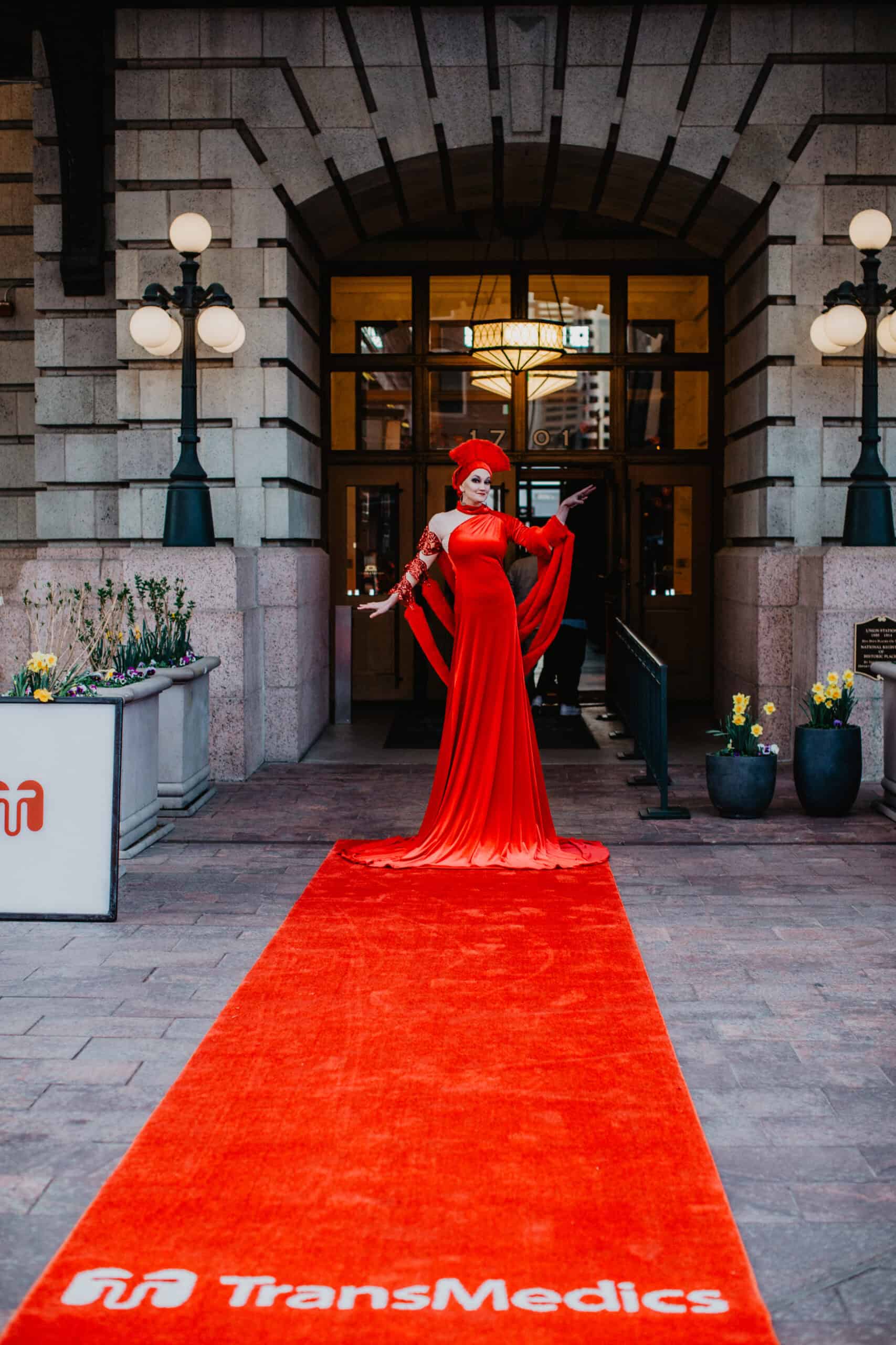 Elegant woman in red gown and mask on red carpet outside historic building.