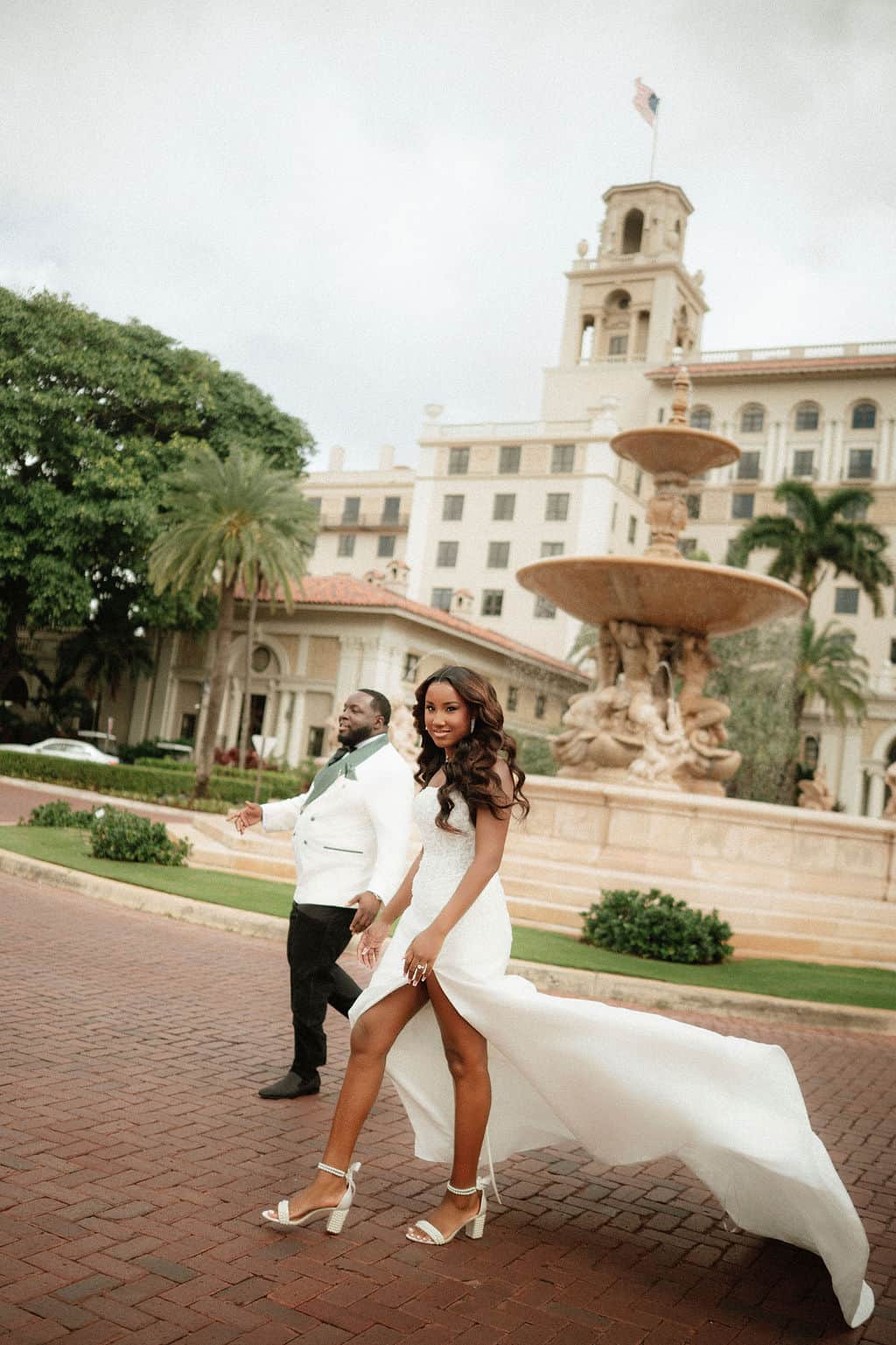 Bride in a white wedding gown with high slit and heels, groom in white suit, historic hotel backdrop.
